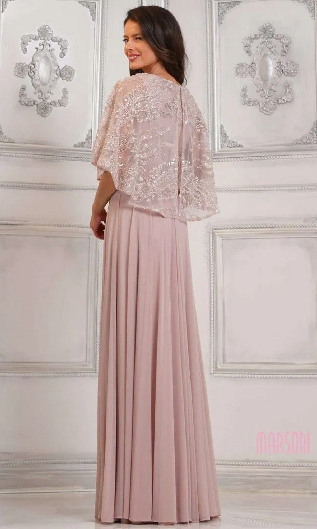 A woman poses in the Marsoni MV1302 long formal dress, featuring beaded details and a sheer embellished capelet, in an ornate, light-colored room.